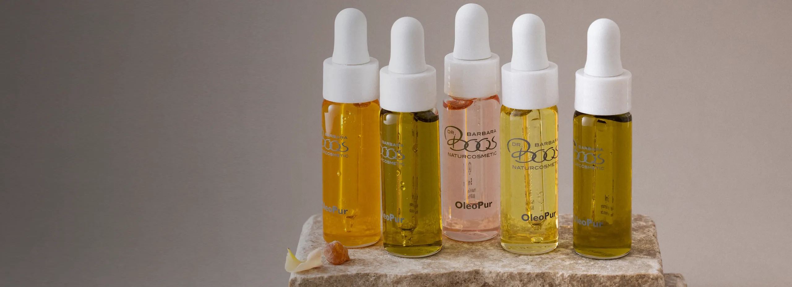 Fünf Glasflaschen mit Pipetten, gefüllt mit verschiedenen Ölen in Gelb- und Rosatönen, stehen auf einem hellen Steinuntergrund. Einige sind mit Etiketten beschriftet. Im Vordergrund liegen kleine Steinstückchen.