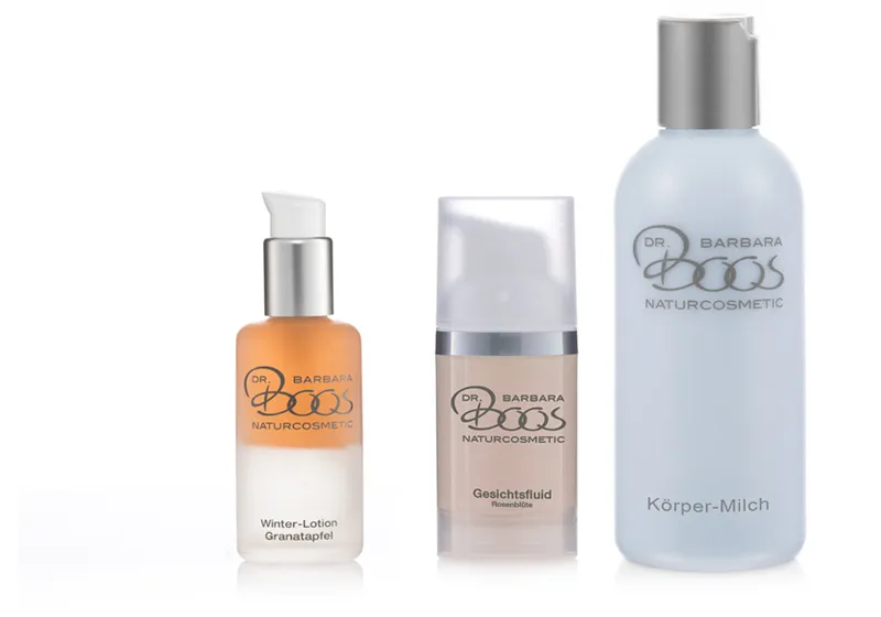 Drei Kosmetikprodukte mit Pump- und Klappverschlüssen: Eine Flasche Winter-Lotion (Granatapfel), ein Spender mit Gesichtsfluid (Rosenblüte) und eine Flasche Körper-Milch. Alle tragen das Logo "Dr. Barbara Boos Naturcosmetic".