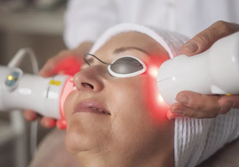 Eine Person mit einer weißen Kappe liegt entspannt, während eine Behandlung mit zwei weißen Geräten mit roten Lichtern im Gesicht durchgeführt wird. Die Person trägt Schutzbrillen, die Augen bedecken.