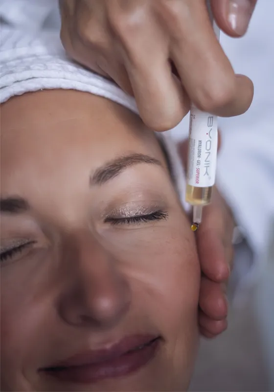 Eine Person mit geschlossenen Augen und einem weißen Kopfhandtuch erhält eine kosmetische Behandlung. Eine Hand trägt ein Serum aus einer kleinen Pipette neben dem Auge auf die Haut auf.