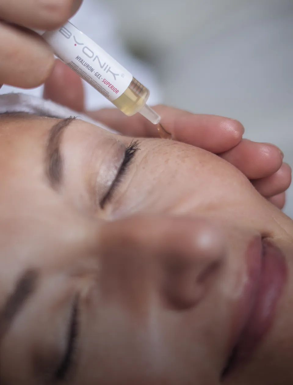 Eine Person mit geschlossenen Augen erhält eine Gesichtsbehandlung. Eine Hand trägt mit einer kleinen Pipette eine Flüssigkeit auf die Haut in der Nähe der Augenbraue der Person auf.