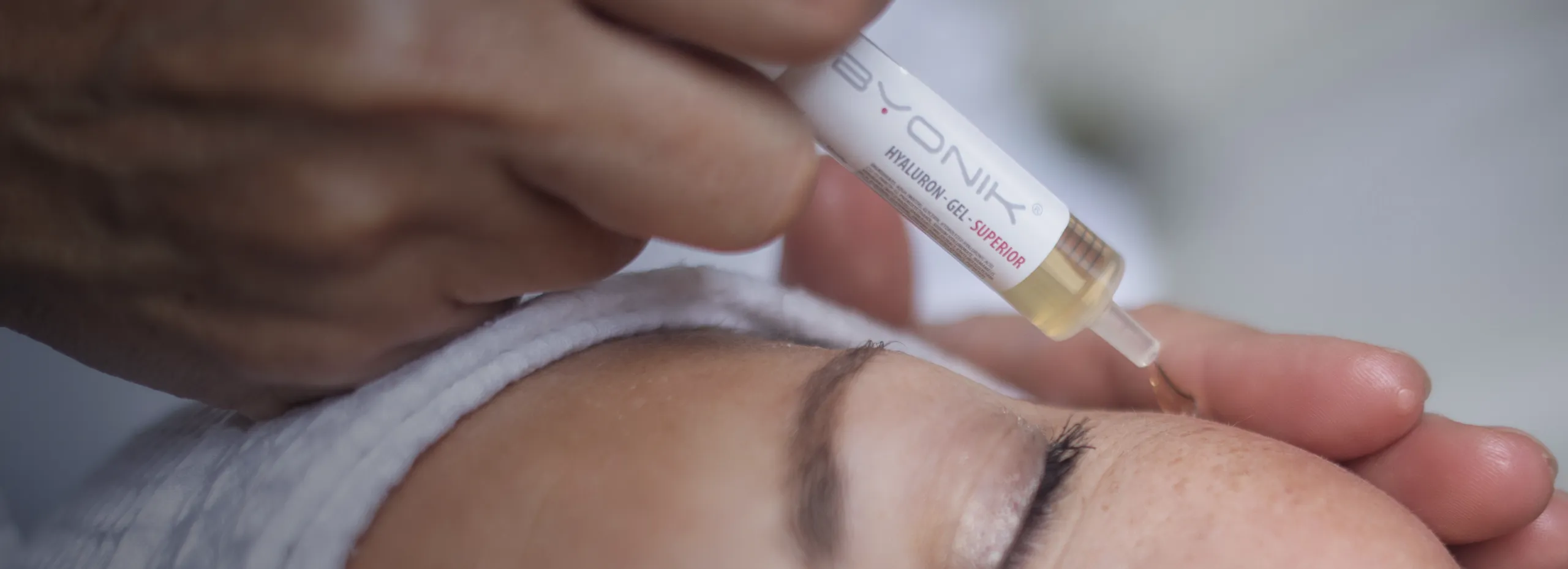 Eine Nahaufnahme zeigt eine Person mit geschlossenem Auge, die ein weißes Stirnband trägt. Ein Hand hält eine Pipette, um eine Flüssigkeit auf die Haut der Person aufzutragen.