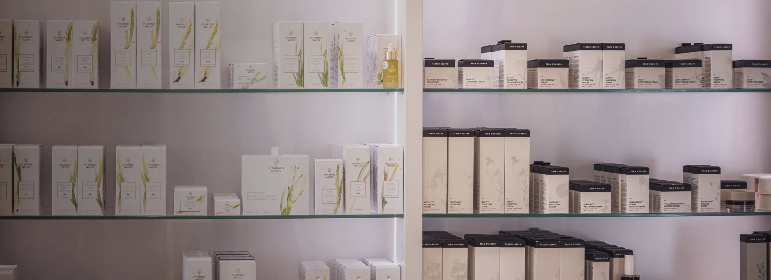 Die Aufnahme zeigt zwei Glasregale gefüllt mit verschiedenen Kosmetikprodukten in weißen und beige Schachteln. Die Produkte sind ordentlich nebeneinander angeordnet, der Hintergrund ist schlicht weiß.