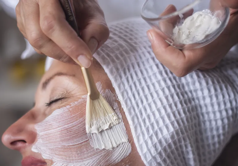 Eine Person mit geschlossenen Augen bekommt eine weiße Gesichtsmaske aufgetragen. Eine Hand hält einen Pinsel, die andere Hand hält eine Schale mit der Maske. Die Person trägt ein weißes Tuch auf dem Kopf.