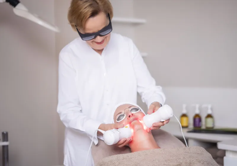 Eine Person liegt auf einer Behandlungsliege und erhält eine Gesichtsbehandlung mit Lichtgeräten. Eine andere Person steht daneben, trägt eine Schutzbrille und führt die Behandlung durch. Im Hintergrund sind verschiedene Flaschen zu sehen.