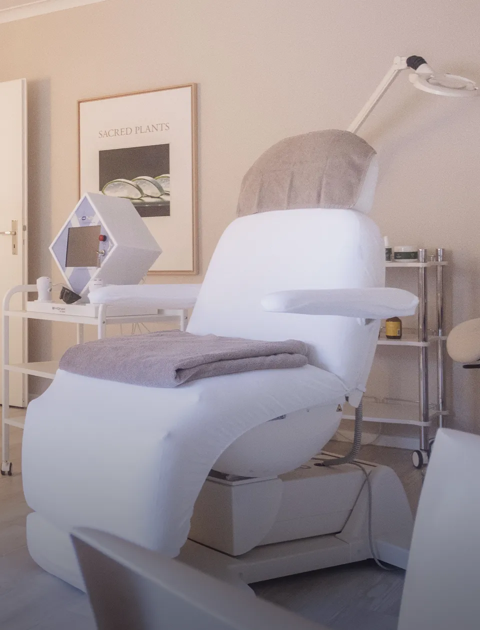 Ein weißer Behandlungsstuhl mit Handtüchern steht in einem Raum. Daneben ist ein Wagen mit einem sechseckigen Gerät und Kosmetikartikeln. An der Wand hängt ein Bild mit der Aufschrift "SACRED PLANTS". Eine Lampe befindet sich über dem Stuhl.