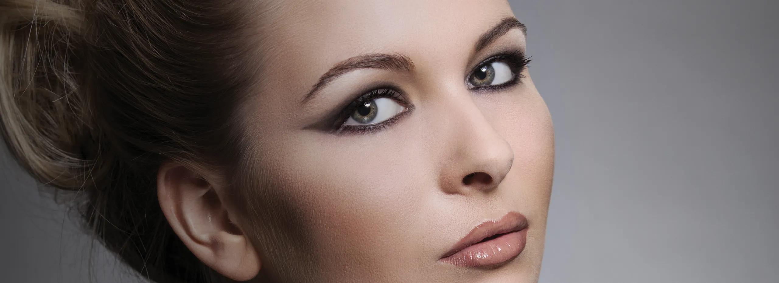 Eine Frau mit hochgestecktem Haar und stark geschminkten Augen blickt leicht seitlich in die Kamera. Der Hintergrund ist grau und schlicht.