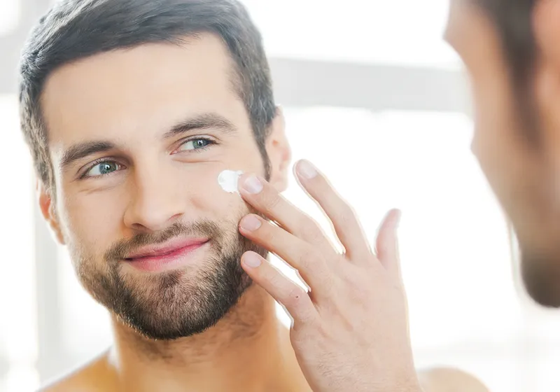 Ein Mann mit Bart trägt Creme auf seine Wange auf. Er lächelt und schaut in einen Spiegel, während er im Badezimmer steht. Die Szene wirkt hell und freundlich.