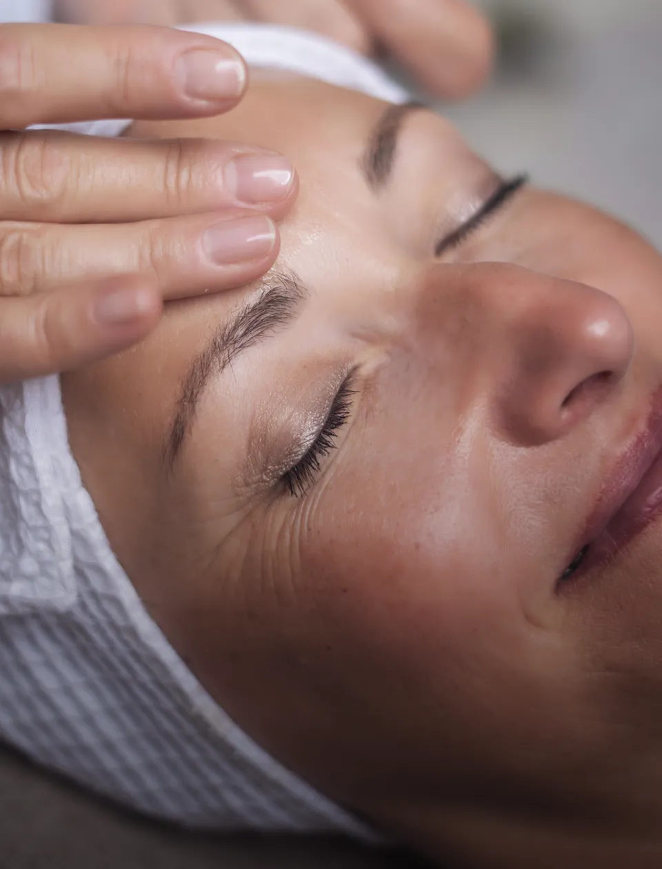Eine Person liegt mit geschlossenen Augen und lächelt leicht. Sie trägt ein weißes Tuch um den Kopf. Hände sind sanft auf die Stirn gelegt, als ob sie eine entspannende Behandlung oder Massage erhält.