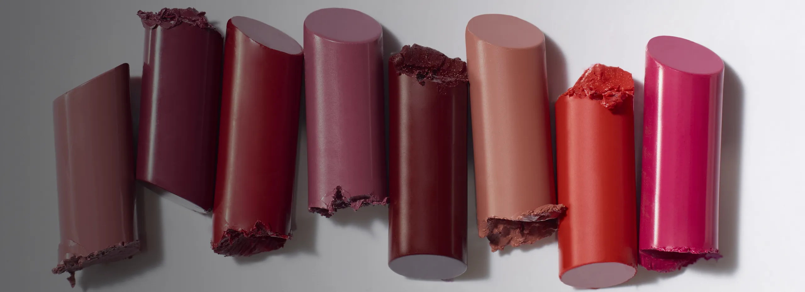 Sieben verschiedene, abgebrochene Lippenstiftstücke in einer Reihe liegen auf einer hellen Oberfläche. Die Farben variieren von dunklem Rot über Lila und Rosa bis zu einem kräftigen Orange und Rot.