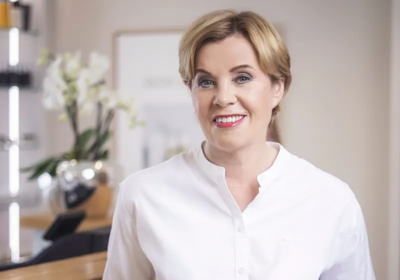 Eine lächelnde Person mittleren Alters mit kurzem, hellbraunem Haar trägt ein weißes Hemd. Im Hintergrund sind ein unscharfes Bild, ein Blumenarrangement und ein Teil eines modernen Raumes zu sehen.