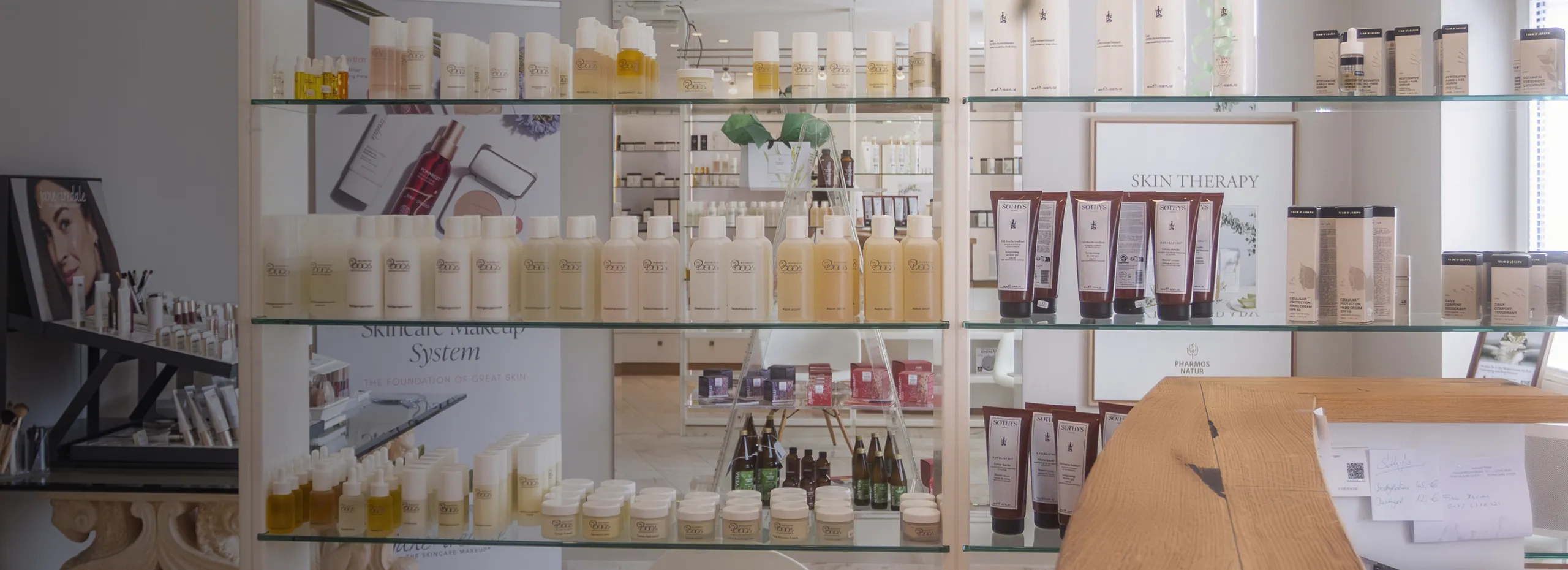 Regale mit verschiedenen Kosmetikprodukten. Auf dem oberen Regal stehen Flaschen mit hellen Flüssigkeiten. Darunter befinden sich Tuben und Flaschen in verschiedenen Formen. Im Hintergrund sind Schilder und weitere Produkte zu sehen.