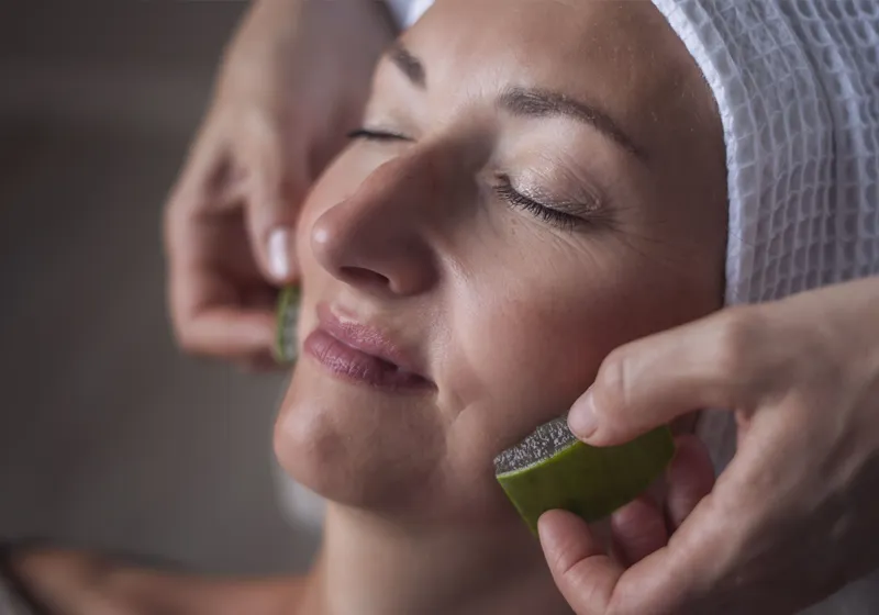 Eine Person liegt entspannt mit geschlossenen Augen. Ein Tuch bedeckt ihr Haar. Zwei Hände halten je ein Stück Luffa-Seife an ihren Wangen. Die Szenerie wirkt wie eine Schönheits- oder Wellnessbehandlung.