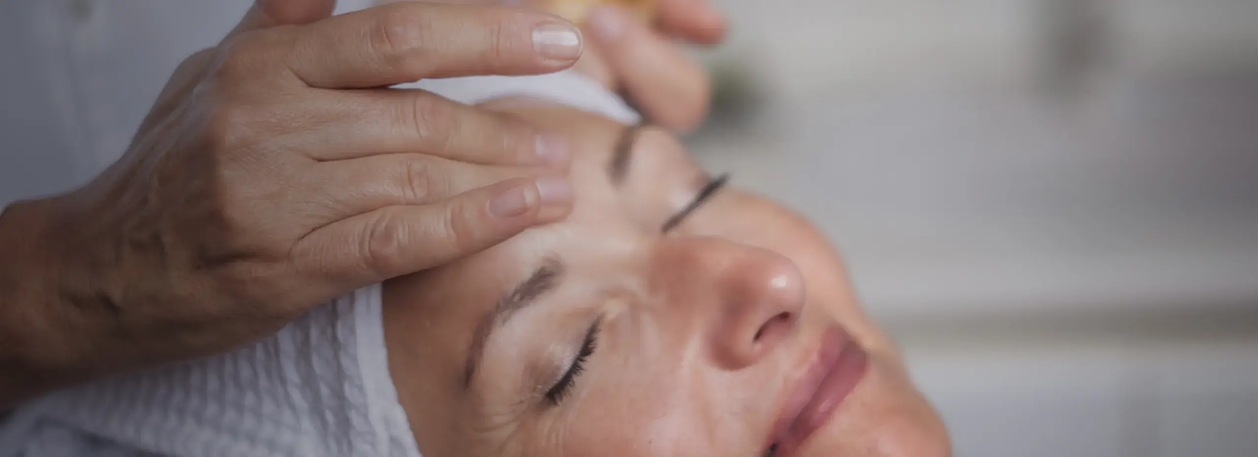 Eine Person liegt mit geschlossenen Augen und entspanntem Gesicht auf einer Liege. Eine andere Hand massiert sanft die Stirn der liegenden Person. Die Atmosphäre wirkt ruhig und wohltuend.