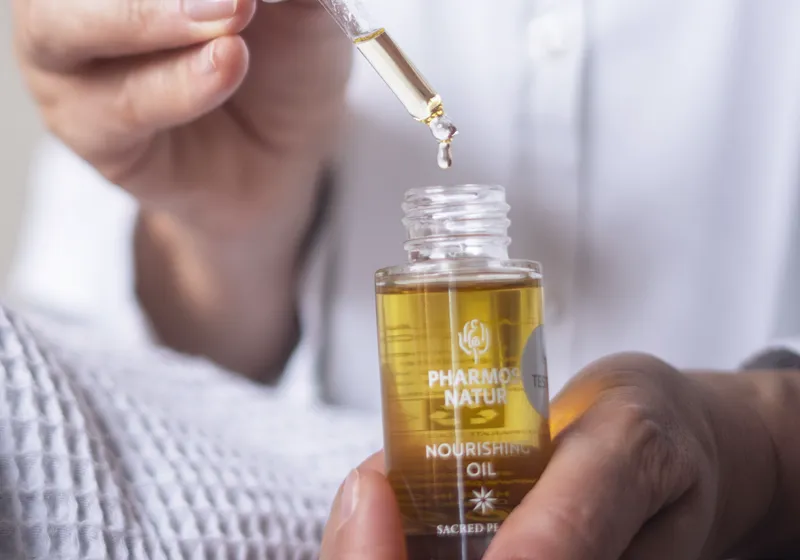 Eine Hand hält eine geöffnete Flasche mit goldenem Öl, während eine andere Hand mit einer Pipette einen Tropfen entnimmt. Das Etikett der Flasche ist teilweise sichtbar. Der Hintergrund ist unscharf.