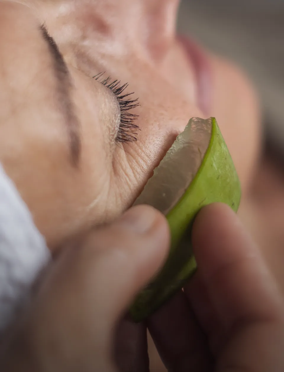 Nahaufnahme einer Person, die ein Stück frischen Aloe-Vera-Gel auf die Haut unter ihrem Auge aufträgt. Die Hand hält das grüne, gelartige Stück vorsichtig an die Gesichtshaut.