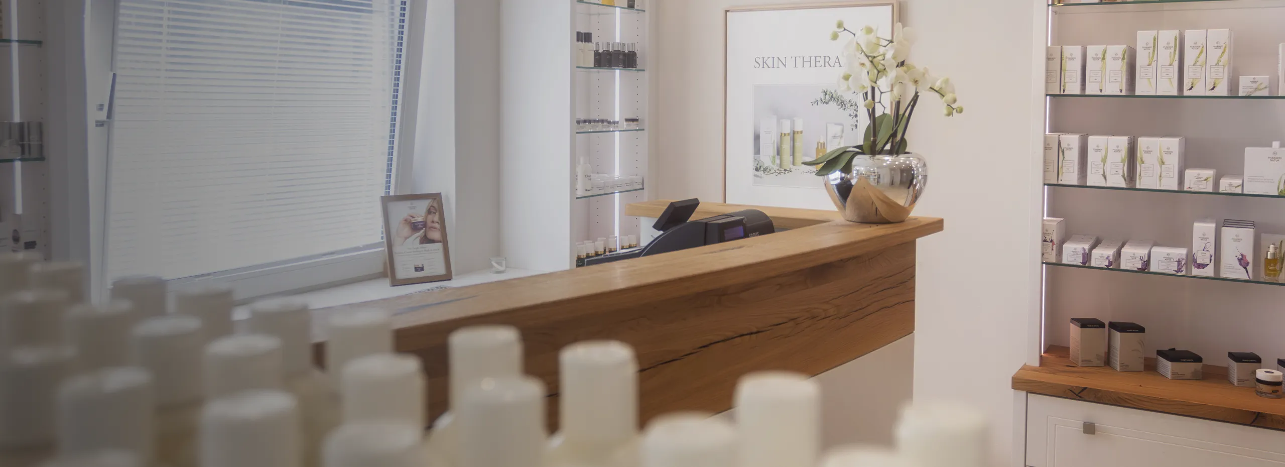Empfangsbereich eines Kosmetikstudios mit Holztheke. Im Hintergrund Regale mit Pflegeprodukten, ein Pflanzenarrangement auf der Theke und ein Bild an der Wand. Links ein Fenster mit Jalousien und einem kleinen Bild.