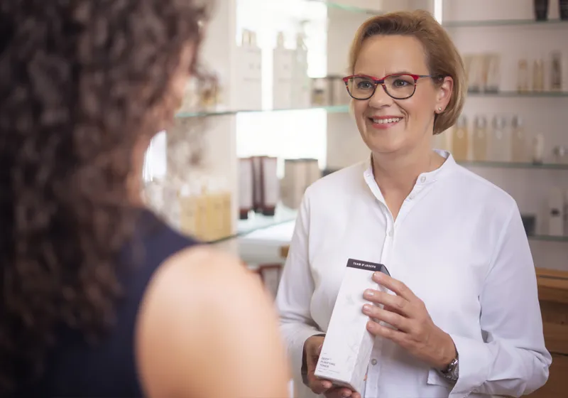 Eine lächelnde Person mit Brille und weißem Hemd steht in einem Geschäft und hält ein Produkt. Im Hintergrund sind Regale mit kosmetischen Produkten zu sehen. Eine andere Person mit lockigem Haar ist von hinten zu sehen.