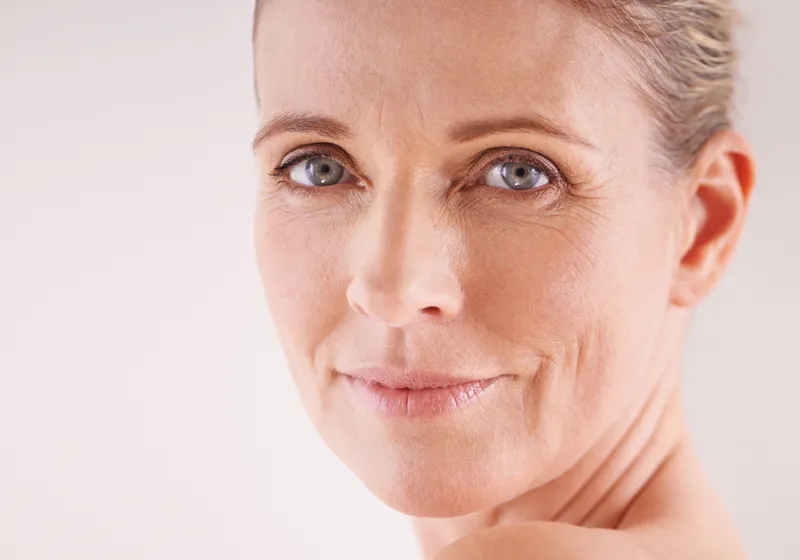 Porträt einer lächelnden Person mittleren Alters mit blondem Haar. Die Person hat blaue Augen und zeigt leichte Lachfalten. Der Hintergrund ist schlicht und hell, was das Gesicht betont.