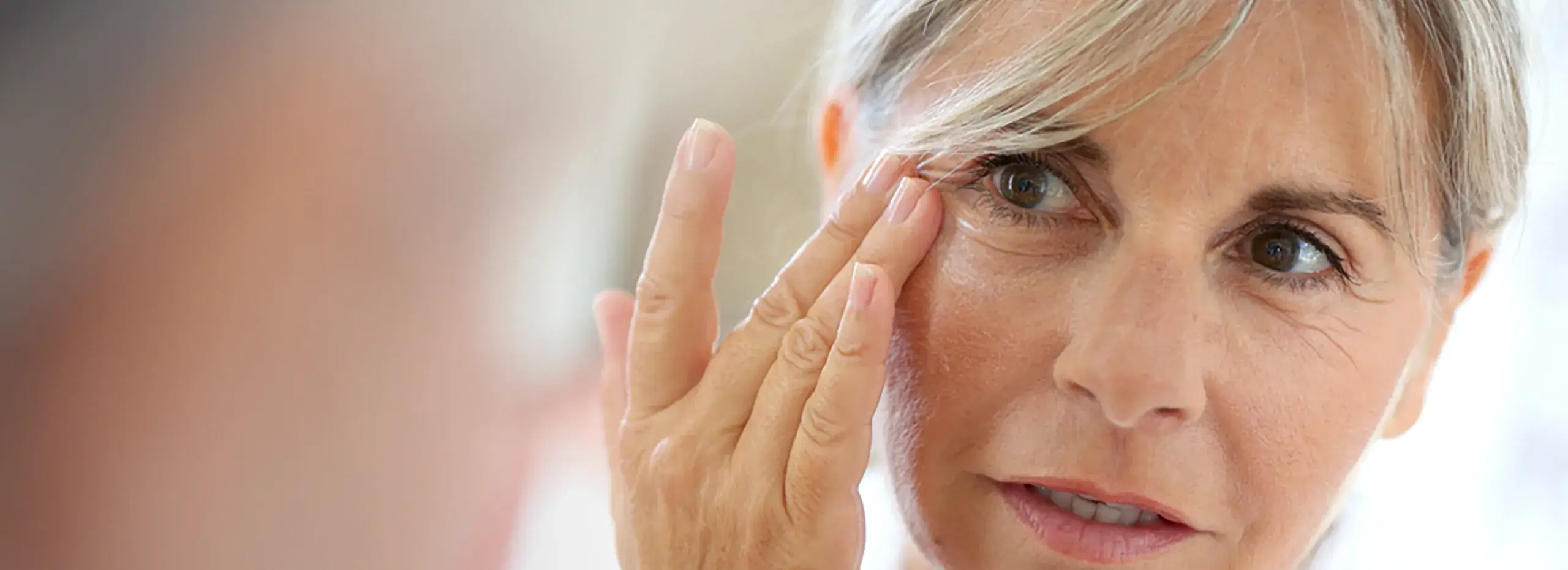 Eine Person mittleren Alters betrachtet ihr Gesicht in einem Spiegel und berührt sanft die Haut um ihre Augen. Sie hat graues Haar und einen konzentrierten Blick. Der Hintergrund ist unscharf.
