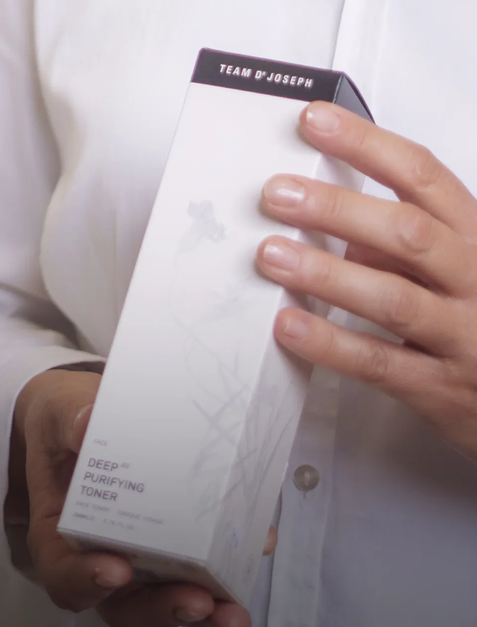 Eine Person in einem weißen Hemd hält eine Box mit der Aufschrift "Deep Purifying Toner" in den Händen. Oben auf der Verpackung steht "Team Dr Joseph". Die Person zeigt die Vorderseite der Box.