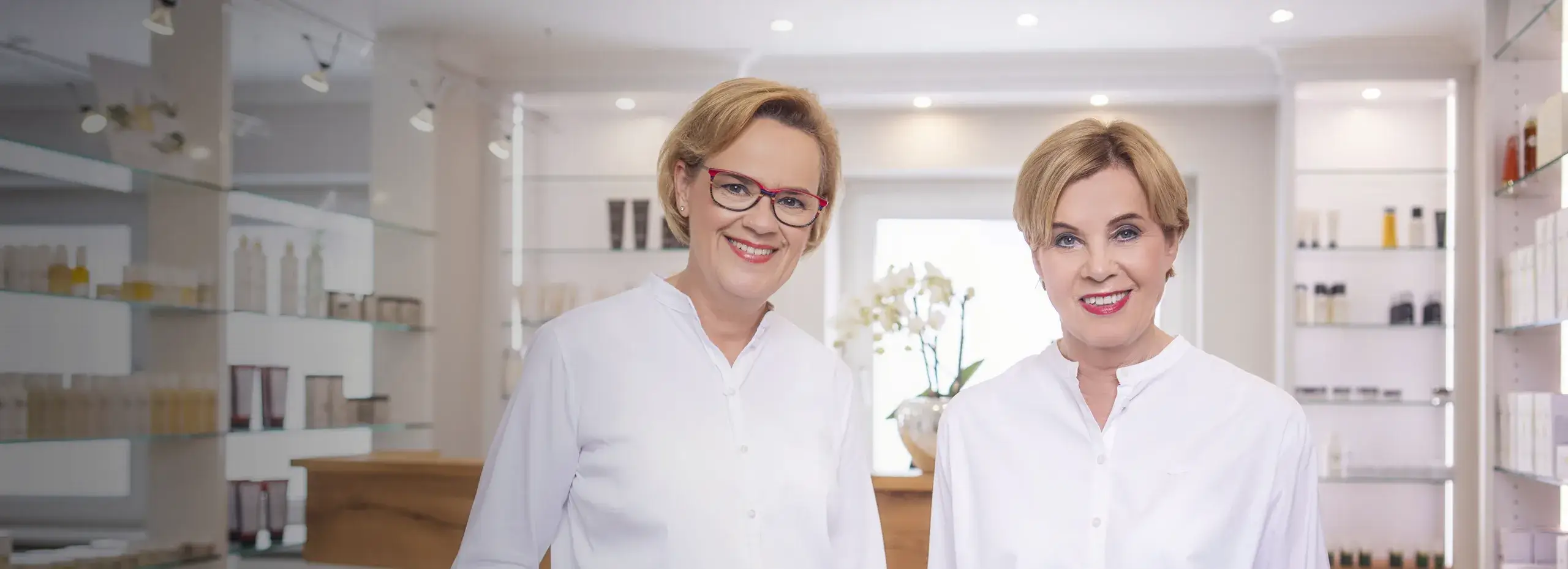 Zwei Personen in weißen Blusen stehen lächelnd in einem hellen Raum, der wie ein Geschäft oder Salon aussieht. Im Hintergrund sind Regale mit verschiedenen Flaschen und Orchideen auf einem Holztisch zu sehen.