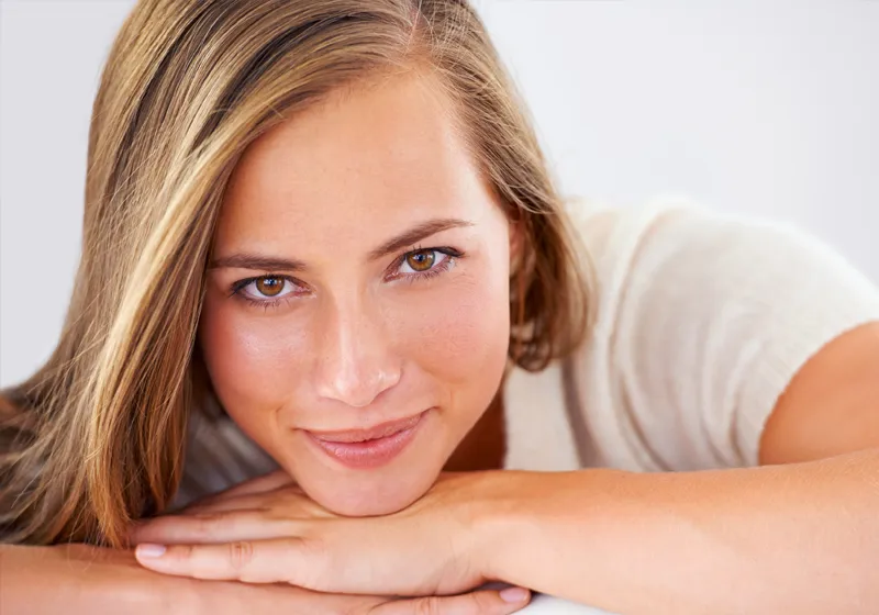Eine Person mit schulterlangem Haar liegt mit dem Kopf auf verschränkten Armen und lächelt direkt in die Kamera. Der Hintergrund ist neutral und hell.