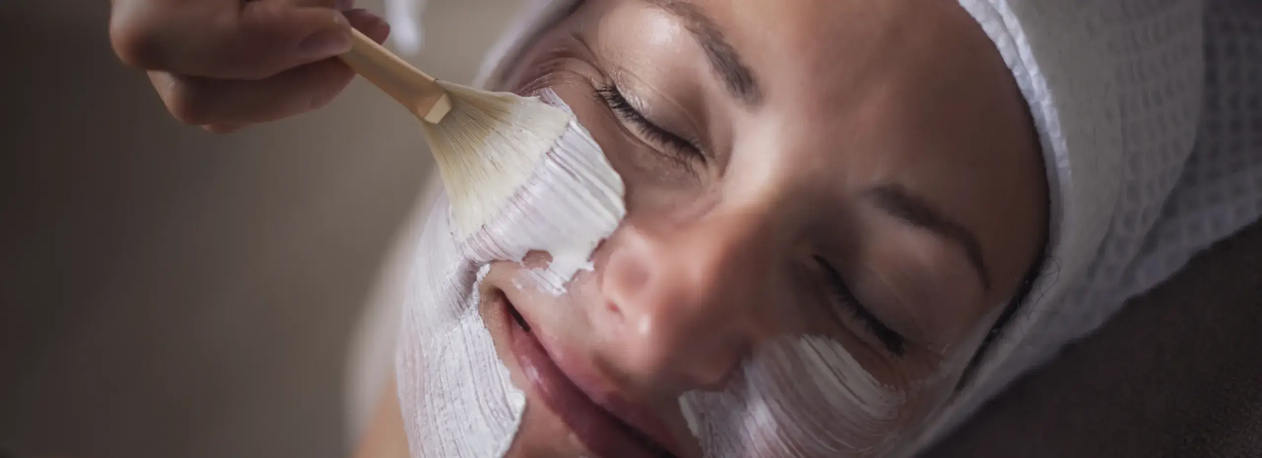 Eine Person mit geschlossenen Augen liegt entspannt und lächelt, während eine Gesichtspackung mit einem Pinsel aufgetragen wird. Sie trägt ein Handtuch um den Kopf.